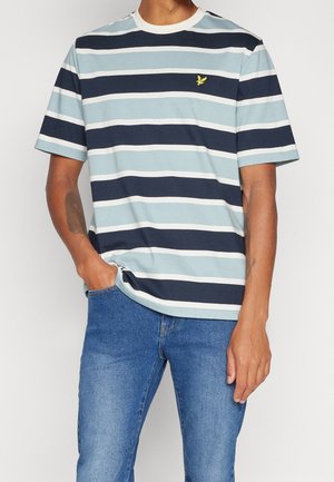 Man draagt een kort mouw gestreept t-shirt in lichtblauw, marineblauw en wit met een klein geel logo, gecombineerd met blauwe spijkerbroek.