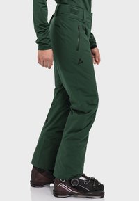 Grüne Skihosen mit geradem Schnitt, ausgestattet mit Reißverschlusstaschen und einem Logo, kombiniert mit braunen Skischuhen. Das Material wirkt strapazierfähig und wasserabweisend.