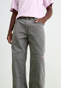 Gray cotton pants with a relaxed fit, featuring seam details and a small logo patch on the leg. Textured fabric with a slightly worn appearance.
