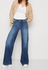 Vaaleansiniset leveälahkeiset farkut, joissa on vaalea pesu, yhdistettynä beigellä raidoitettuun bleiseriin ja valkoiseen V-pääntie t-paitaan, jossa on metallinvärinen teksti.