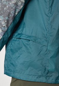 Teal jakke med teksturert finish, med grå mønstret stoff på ermene og en sømløs sidelomme for oppbevaring.