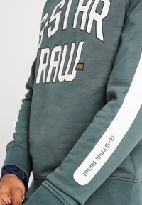 Zelená mikina s výrazným bílým nápisem "G-STAR RAW." Obsahuje bílé pruhy na rukávech a malou nášivku značky. Měkká látka, žebrované manžety.