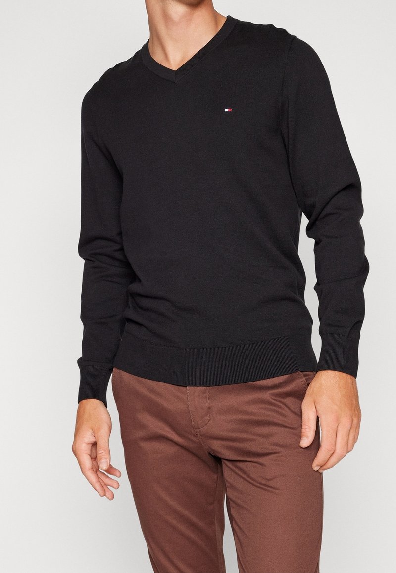 Maglione nero a maglia con scollo a V, polsini e orlo a coste; piccolo logo sul lato sinistro del petto; indossato con pantaloni chinos marroni.