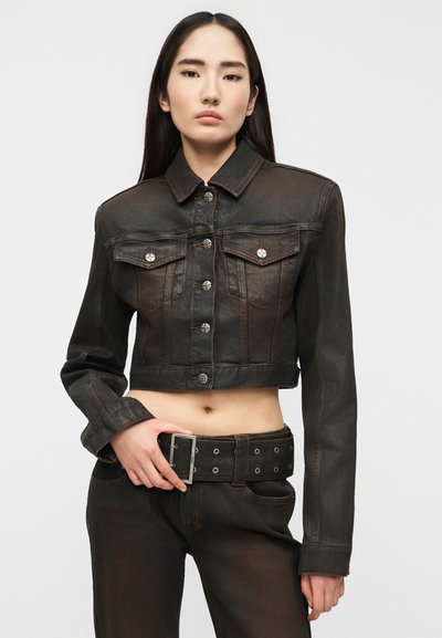 Νεαρή γυναίκα που φοράει μαύρο τζιν μπουφάν cropped και μαύρο τζιν ψηλόμεσο με φαρδιά μαύρη ζώνη, στέκεται μπροστά σε απλό φόντο.