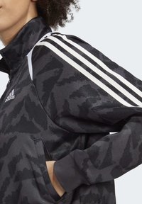 Black athletic jacket with a camouflage pattern, white three-stripe accents, and a high collar. Features ribbed cuffs and side pockets.