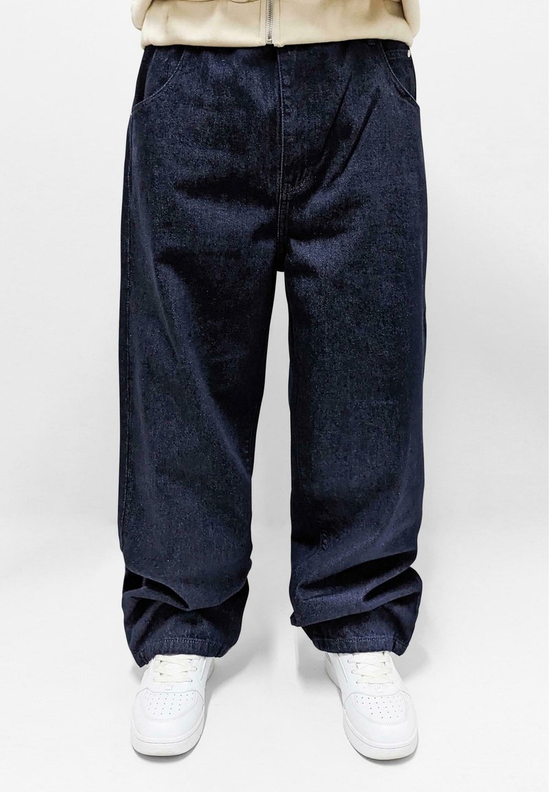 Donkerblauwe denim broek met een losse pasvorm, voorzien van zijzakken en een taps toelopende zoom. Gecombineerd met witte sneakers voor contrast.