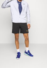 Chaqueta Nike de color gris claro con cierre de cremallera, camiseta azul debajo, pantalones cortos negros con logo, calcetines blancos y zapatillas deportivas azules con detalles.