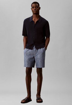 Man staat, draagt een zwart kortarmig overhemd met knopen, lichtblauwe korte broek en donkerbruine sandalen tegen een effen grijze achtergrond.