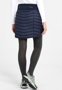 Marinblå vadderad isolerad kjol med midjeband som har Columbia-logotypen, kombinerad med grå leggings och grå/gröna sportskor.