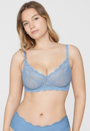 Kobieta z długimi włosami, ubrana w pasujący jasnoniebieski koronkowy biustonosz i bieliznę, stojąca na jednolitym jasnym tle.