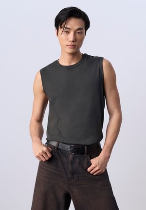 Junger Mann mit schwarzen Haaren, der ein ärmelloses schwarzes Shirt in hoch taillierte schwarze Jeans mit schwarzem Gürtel steckt, Hände in den Taschen.