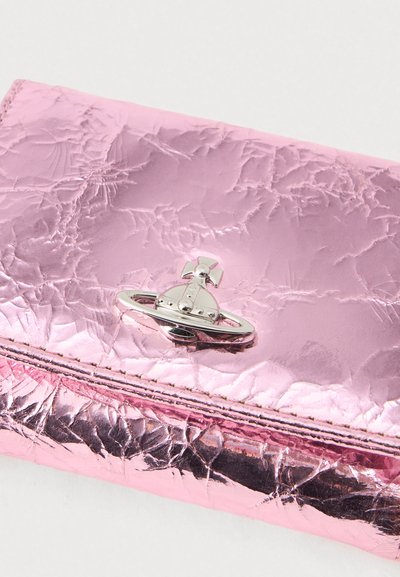 Rózsaszín fémes textúrájú pénztárca, ezüst gömb alakú logóval, amelyen kereszttel és gyűrűrészlettel díszített, világos háttérrel.