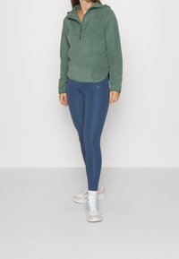 Pullover in pile verde con zip a metà, abbinato a leggins blu e scarpe da ginnastica pastello. Sullo sfondo neutro.