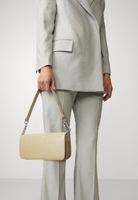 Beige keinonahka käsilaukku, jossa on teksturoitu krokotiilikuosi, hopeiset yksityiskohdat ja irrotettava hihna, jota pitää henkilö vaaleanharmaassa puvussa.