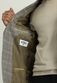 Grijs geruite blazer met een gladde olijfgroene voering, voorzien van een gelabelde binnenzak en een tan accent. De buitenkant toont een gestructureerd ontwerp.