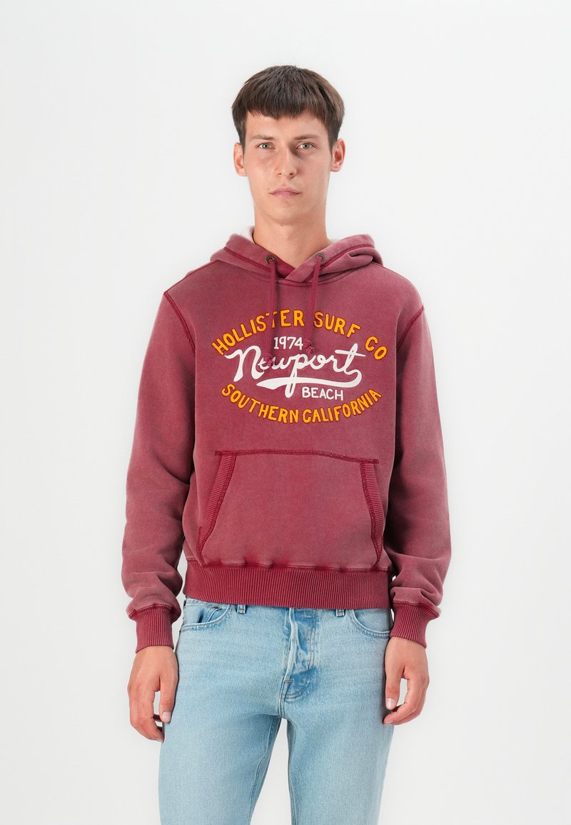 Sudadera marrón de algodón, con un bolsillo canguro frontal, cordones y texto impreso en amarillo y blanco para la marca.