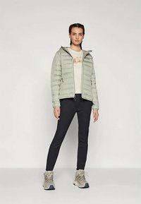 Frau steht vor einem schlichten weißen Hintergrund und trägt eine hellgrüne Pufferjacke, ein beiges T-Shirt mit Grafik, schwarze Hosen und beiges Wanderstiefel.