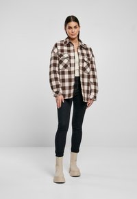 Chaqueta de camisa a cuadros marrón y crema con dos bolsillos en el pecho, cierre de botones y mangas largas, combinada con jeans negros y botas de tobillo beige.