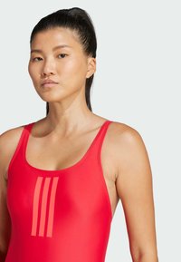 Red swimwear with a smooth, stretchy texture. Features wide shoulder straps and three vertical stripes in a lighter shade on the front.