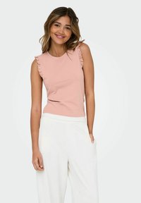 Rosa ärmlös topp med volangdetaljer i spets på axlarna, gjord av mjukt tyg, i kombination med vita vida byxor.