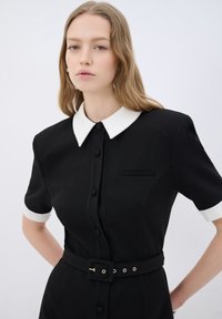 Schwarzes Kleid mit kurzen Ärmeln und weißen Manschetten, ausgestattet mit einem weißen Kragen, vorderen Knöpfen und einer taillierten Taille mit goldenen Akzenten.