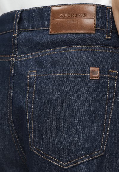 Σκούρα μπλε τζιν με ραφές σε ταμπέ χρώμα, δερμάτινο πλαίσιο με την επιγραφή "WEEKEND Max Mara" και ένα μικρό έμβλημα πεταλούδας στην πίσω τσέπη.
