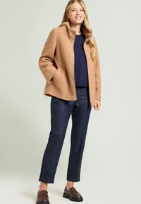 Giacca di fleece beige con colletto alto, chiusura a zip frontale e tasche laterali, abbinata a pantaloni a righe blu marino e mocassini marroni.