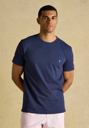Ung mand stående med hænderne bag ryggen iført en mørkeblå t-shirt og lyserøde bukser mod en neutral baggrund.