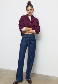 Pleteni maro pulover z visokim ovratnikom in gumbi, vparen z visokimi temno modrimi traper hlačami v zvonastem kroju in rjavimi usnjenimi čevlji.