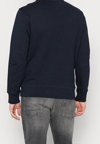 Donkerblauwe sweat met ronde hals, geribbelde manchetten en zoom. Heeft een gladde textuur en minimalistisch ontwerp. Draagt met grijze spijkerbroek.