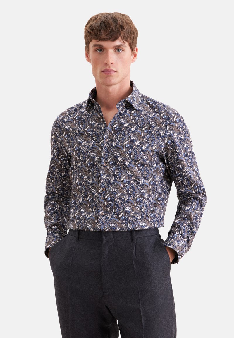 Jeune homme portant une chemise boutonnée à manches longues avec un motif floral paisley bleu et marron et un pantalon gris foncé, les mains dans les poches.