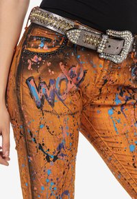 Pantalon en denim orange avec des éclaboussures de peinture multicolores. Il est doté d'une ceinture décorative avec des clous en argent et une grande boucle ornée.