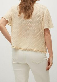 Beige short-sleeve textured blouse with pom-pom trim paired with off-white fitted pants, shown from the back.