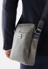Borsa a spalla grigia in tessuto con superficie texture, caratterizzata da un accento in pelle, tracolla regolabile e una patch con logo sul davanti.