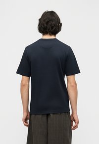 Άτομο με κοντά κυματιστά μαύρα μαλλιά, που φοράει μια απλή σκούρα μπλε μπλούζα και φαρδιά σκούρα καφέ παντελόνια, με την πλάτη προς τα εμπρός σε ένα απλό φόντο.