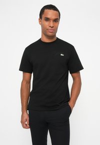 T-shirt en coton noir à manches courtes, col rond, avec un petit logo vert brodé sur la poitrine. Design simple et classique.