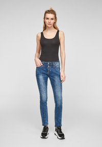 Schwarzes Tanktop, blaue schmal geschnittene Jeans mit ausgewaschenem Finish, fünf Taschen, Knopfverschluss, getragen mit schwarzen Turnschuhen. Einfaches, lässiges Outfit.