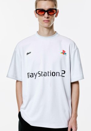 Bijela oversized majica sa svijetloplavim ovratnikom, s crnim natpisom "PlayStation 2" i šarenim PlayStation logom na prsima. Crne hlače i crvene sunčane naočale.