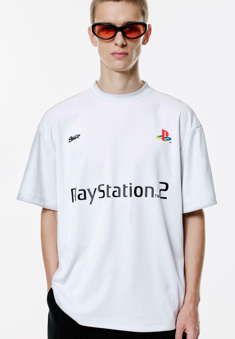Bijela oversized majica sa svijetloplavim ovratnikom, s crnim natpisom "PlayStation 2" i šarenim PlayStation logom na prsima. Crne hlače i crvene sunčane naočale.