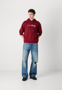 Maroon hoodie with drawstrings and front pocket, featuring white and blue "Calvin Klein New York" logo; paired with distressed light blue wide-leg jeans.