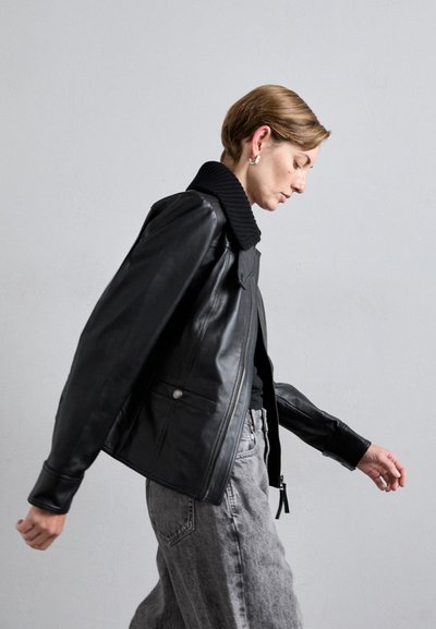 Black leather bomber jacket with a ribbed collar, zip front, and side pockets. Worn over a black top and paired with gray denim.