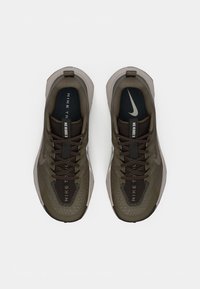Olīvzaļa un melna Nike skriešanas apavu pāris ar saites, balta Nike swoosh un teksts "NIKE TRAIL" uz iekšējo zolīšu.