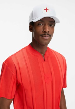 Uomo che indossa una maglietta a righe rosse e un cappellino bianco con un emblema a croce rossa, che guarda direttamente la fotocamera su uno sfondo semplice.