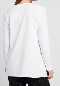 Top bianco a maniche lunghe con texture a coste, scollatura rotonda e spacchi laterali; vestibilità casual, abbinato a pantaloni neri.