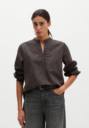 Vrouw draagt een blouse met lange mouwen met patroon en opgerolde mouwen en een hoog getailleerde donkergrijze spijkerbroek, staand met één hand in de zak.