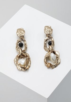 Pendientes de tono dorado con un diseño orgánico texturizado, que presentan una perla negra y una perla en forma de lágrima blanca suspendida dentro de anillos circulares.