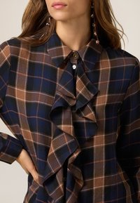 Camisa de cuadros marrón y azul marino con un frontal con volantes, tapeta de botones y un cuello clásico. Fabricada con un tejido suave de textura lisa.
