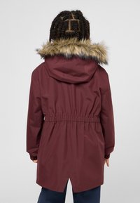 Bordeauxfarbener Parka mit Kunstfell gefütterter Kapuze, tailliertem Schnitt und elastischen Bündchen. Verfügt über einen ventilierten Rückensaum und glattes, wasserabweisendes Material.