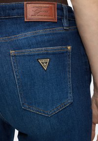 Μπλε τζιν τζιν με το τρίγωνο λογότυπο Guess στην πίσω τσέπη και δερμάτινη ετικέτα στη μέση με την ένδειξη "Guess Hope Never Give Up."