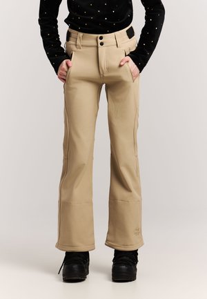 Personne portant un pantalon beige à jambes larges avec des poches avant et des bottes noires, les mains dans les poches, debout devant un fond blanc.